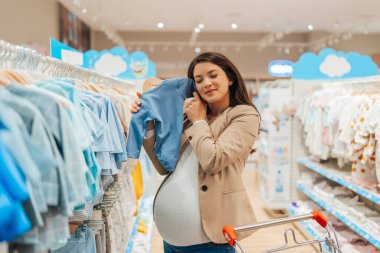 Beautiful young pregnant mother choosing and buying colorful clothes and appliances for her new incoming baby in. Child shop or store concept.