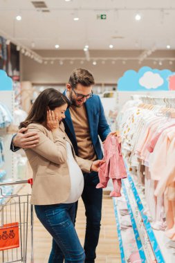 Attractive middle age couple enjoying in buying clothes and appliances for their new baby. Heterosexual couple in baby shop or store. Expecting baby concept.