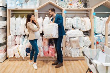 Attractive middle age couple enjoying in buying clothes and appliances for their new baby. Heterosexual couple in baby shop or store. Expecting baby concept.