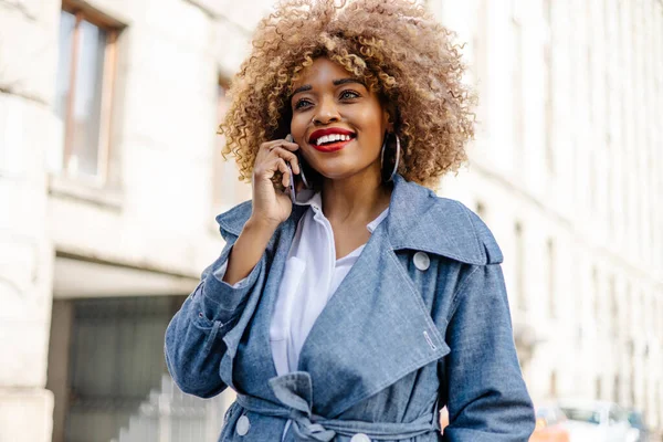 Beautiful black woman walks down the street and uses smart phone for communication. She is happy and smiled. Bright sunny day.