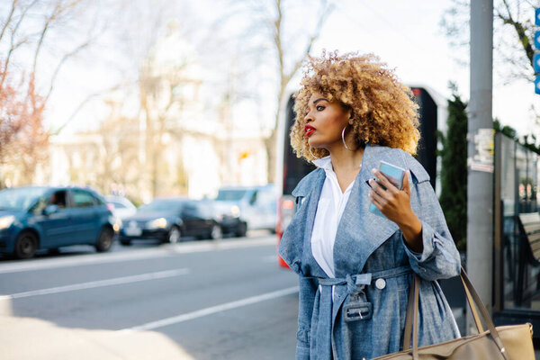 Beautiful black woman walks down the street and uses smart phone for communication and calling for taxi with raised arm. She is happy and smiled. Bright sunny day.