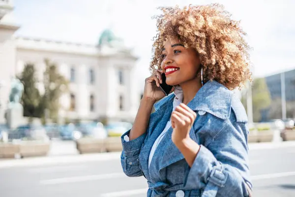 美丽的黑人妇女走在街上 用智能手机进行交流 她很高兴 阳光灿烂的日子 — 图库照片