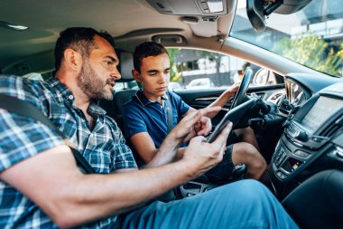 Öğrencisiyle arabada oturan bir sürücü eğitmeni. Ona temel direksiyon ve trafik kurallarını açıkla. Eğitim amaçlı olarak elektronik tablet kullanıyor. İçeriden görüntüle.