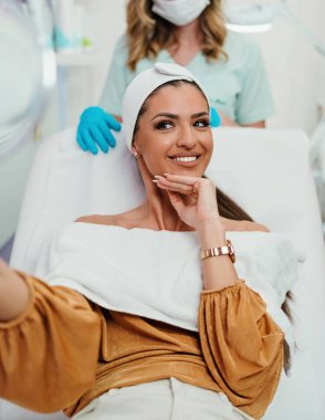 Güzel ve mutlu genç bir kadın tıbbi sandalyede oturup aynaya bakıyor. Hyaluronik asit doldurucu ya da botulinyum toksin iğneleriyle başarılı bir güzellik tedavisinden sonra tatmin oluyor..