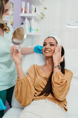 Güzel ve mutlu genç bir kadın tıbbi sandalyede oturup aynaya bakıyor. Hyaluronik asit doldurucu ya da botulinyum toksin iğneleriyle başarılı bir güzellik tedavisinden sonra tatmin oluyor..