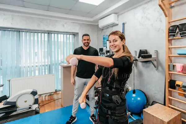 Professional physiotherapist working with beautiful young woman in EMS suit during special electro stimulation training or treatment. Modern and brightly lit studio.