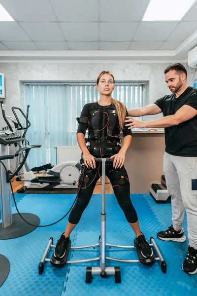 Professional physiotherapist working with beautiful young woman in EMS suit during special electro stimulation training or treatment. Modern and brightly lit studio.