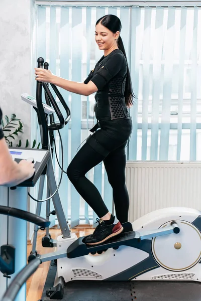 Professional physiotherapist working with beautiful young woman in EMS suit during special electro stimulation training on a stepper or cardio climber. Modern and brightly lit studio.