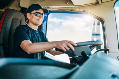 Yakışıklı, şapkalı ve kamyonetini süren bir erkek kamyon şoförü. Profesyonel ulaşım ve kamyon şoförleri kavramı. Yan görünüm.