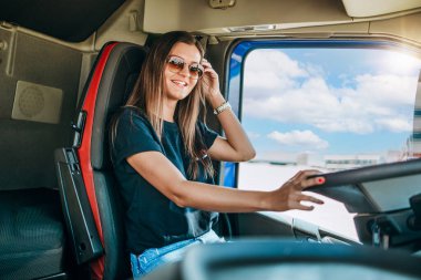 Portrait of beautiful young woman professional truck driver sitting in a big truck, looking at camera and smiling. Inside of vehicle. People and transportation concept.