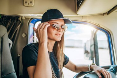 Portrait of beautiful young woman professional truck driver sitting in a big truck, looking at camera and smiling. Inside of vehicle. People and transportation concept.