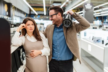 Beautiful and happy middle age couple buying consumer tech products in modern home appliances store. They are choosing home theater and TV devices. People and consumerism concept.