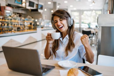 Güzel ve mutlu genç bir kadın fırında ya da fast food 'da oturup lezzetli börekler yiyor. Ayrıca laptopunu kullanıyor ve kulaklıkla müzik dinliyor.. 
