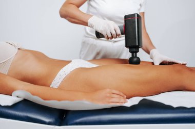 Young sporty caucasian woman having body massage using percussion pistol at beauty relax center. Electric massager pistol for special muscle treatment. Sports and pain recovery concept.