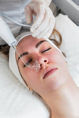 Genç bir kadın yüksek frekanslı değnekle yüz gerdirme tedavisi görüyor. Bu modern tedavi akıma neden olan bakterileri öldürmek, iltihabı azaltmak ve cilt rengini ve dokusunu iyileştirmek için elektrik akımı kullanır..