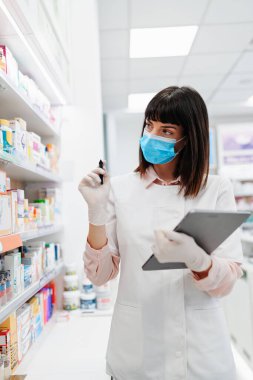 Koruyucu maskeli güzel bir eczacı eczanede çalışıyor ve stok alımı yapıyor. Pozitif sağlık çalışanı portresi ya da iş yerinde çalışan bir kimyager..