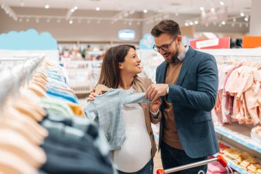 Attractive middle age couple enjoying in buying clothes and appliances for their new baby. Heterosexual couple in baby shop or store. Expecting baby concept.