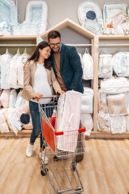 Attractive middle age couple enjoying in buying clothes and appliances for their new baby. Heterosexual couple in baby shop or store. Expecting baby concept.