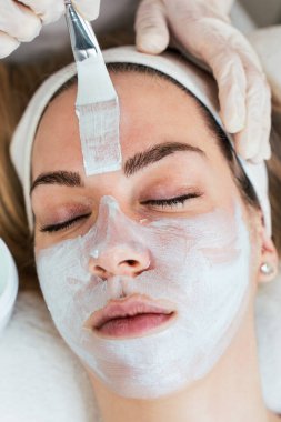 Güzel ve çekici genç bir kadın bitkisel kil maskeyle profesyonel güzellik ya da cilt bakımı görüyor. Modern ve popüler yüz bakımı kavramı. Spa salonu. Yüksek açı görünümü.