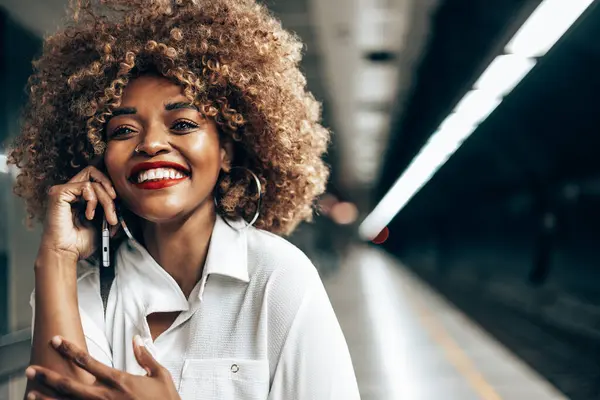 地下鉄の駅に立って美しいファッショナブルな黒人女性 彼女は満足していると彼女のスマートフォン上の誰かと話している 公共交通機関と都市生活の概念 ストック画像