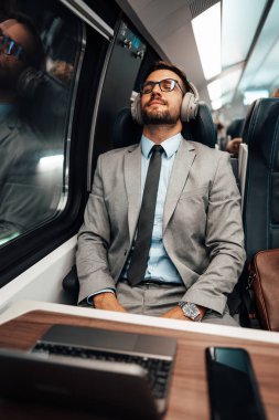 Hızlı tren ya da metro ile seyahat ederken yorgun ve yakışıklı iş adamı dinleniyor. O, eğlence ve müzik dinlemek için dizüstü bilgisayar ve kablosuz kulaklık kullanıyor. Modern ve hızlı seyahat kavramı.