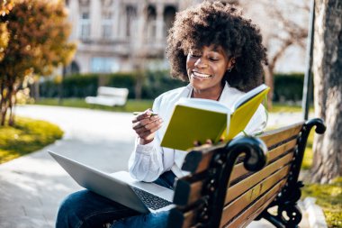 Güzel ve ciddi siyahi kız öğrenci fakülte sınavına çalışırken kampüste dışarıda eğleniyor. O, parkta bankta oturuyor ve dizüstü bilgisayarında bir şey okuyor.. 
