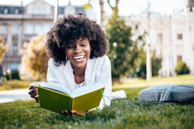 Güzel ve ciddi siyahi kız öğrenci fakülte sınavına çalışırken kampüste eğleniyor. Çimlerin üzerinde uzanıyor ve bir şeyler okuyor. Arka plandaki eski bina.