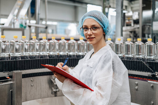 Женщина-работница в защитной рабочей одежде, работающая на научно-исследовательском и производственном заводе медикаментов и проверяющая канистры с дистиллированной водой перед отгрузкой. Контроль качества. 