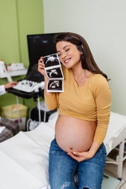 Beautiful and pregnant woman is happy after pregnancy medical check up. She is holding and looking at ultrasound scan of her baby. Modern pregnancy healthcare concept. 