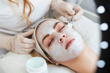 Güzel ve çekici genç bir kadın bitkisel kil maskeyle profesyonel güzellik ya da cilt bakımı görüyor. Modern ve popüler yüz bakımı kavramı. Spa salonu. Yüksek açı görünümü.