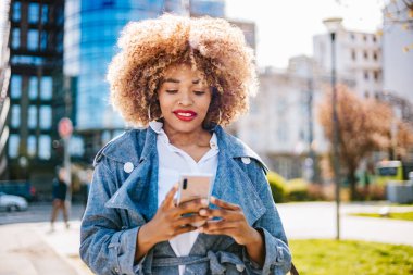 Güzel siyahi kadın sokakta yürüyor ve mesajlaşmak ve iletişim kurmak için akıllı telefon kullanıyor. O mutlu ve gülümsedi. Parlak güneşli bir gün.