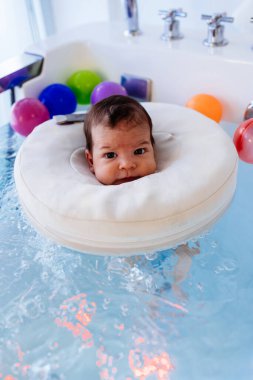 Küçük bir çocuk ya da bebek yüzüyor ve havuzda fizik tedavi seansı sırasında iyi vakit geçiriyor. Bebek kameraya bakıyor ve gülümsüyor..