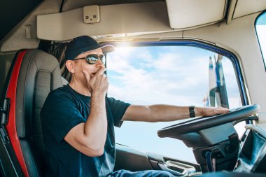 Yakışıklı, şapkalı ve kamyonetini süren bir erkek kamyon şoförü. Profesyonel ulaşım ve kamyon şoförleri kavramı. Yan görünüm.