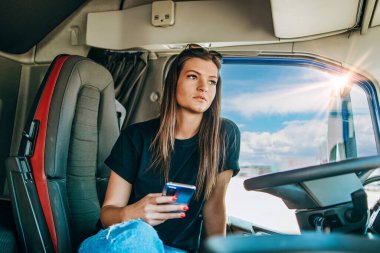Profesyonel kamyon şoförünün uzun bir yolculuktan sonra oturup dinlendiği güzel bir kadın portresi. O, akıllı telefonunu kullanıyor. Aracın içinde. İnsanlar ve ulaşım kavramı.
