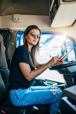 Portrait of beautiful young woman professional truck driver sitting in a big truck, looking at camera and smiling. Inside of vehicle. People and transportation concept.