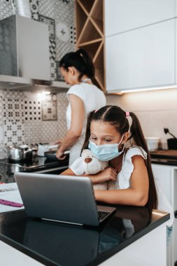 Covid-19 salgını sırasında küçük bir kız e-öğrenim seansı yapıyor ya da karantina altında. Anne mutfakta çalışıyor ve arka planda öğle yemeği hazırlıyor.. 