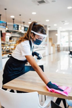 Fırında ya da fast food restoranında çalışan güzel bir kadın. Coronavirüs salgınına karşı masaları temizleyip dezenfekte ediyor. Koruyucu maske, eldiven ve yüz maskesi takıyor..