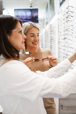 Modern optik mağazada gözlük çerçevesi seçen güzel ve modaya uygun bir kadın. Kadın satıcı uzmanı doğru karar vermesine yardımcı oluyor..