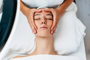 Güzel, rahat, genç bir kadın gözleri kapalı uzanmış bir kaplıca salonunda yüz masajının keyfini çıkarıyor. Bayan müşteri cilt bakımı tedavisinden zevk alıyor. Sağlık ve güzellik kavramı.