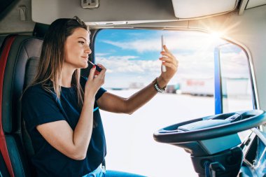 Portrait of beautiful young woman professional truck driver sitting and resting after long drive. She is using her smart phone for online communication. Inside of vehicle. People and transportation concept.