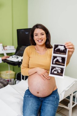 Beautiful and pregnant woman is happy after pregnancy medical check up. She is holding and looking at ultrasound scan of her baby and looking at camera. Modern pregnancy healthcare concept.