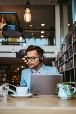 Orta yaşlı yakışıklı adam modern kitapçıda oturuyor, kulaklıkla müzik dinlerken dizüstü bilgisayarını ya da bilgisayarını okuyor ve kullanıyor..