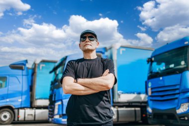 Şapkalı ve güneş gözlüklü profesyonel kamyon şoförü büyük ve modern bir kamyonun önünde duruyor. Parlak güneşli bir gün. İnsanlar ve ulaşım kavramı.