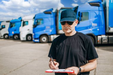 Şapkalı ve güneş gözlüklü profesyonel kamyon şoförü büyük ve modern bir kamyonun önünde duruyor. Parlak güneşli bir gün. İnsanlar ve ulaşım kavramı.