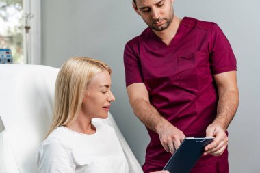 Güzel ve mutlu genç bir kadın tıbbi sandalyede oturup aynaya bakıyor. Hyaluronik asit doldurucu ya da botulinyum toksin iğneleriyle başarılı bir güzellik tedavisinden sonra tatmin oluyor..