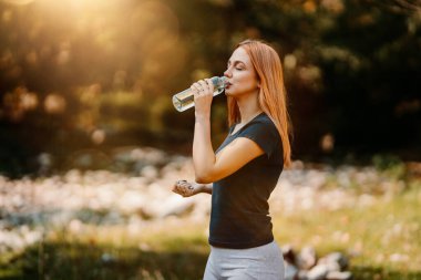 Güzel kızıl saçlı kadın ormanda yürüyor ve bir şişe içme suyuyla koşuyor. Doğa konseptinde sağlıklı yaşam tarzı ve spor.