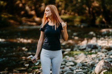 Güzel kızıl saçlı kadın ormanda yürüyor ve bir şişe içme suyuyla koşuyor. Doğa konseptinde sağlıklı yaşam tarzı ve spor.