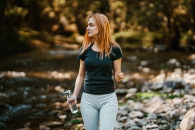 Güzel, genç, kızıl saçlı kadın koşudan sonra rahatlıyor. Nehir kenarında yürür ve doğanın tadını çıkarır. Doğa konseptinde sağlıklı yaşam tarzı ve spor.