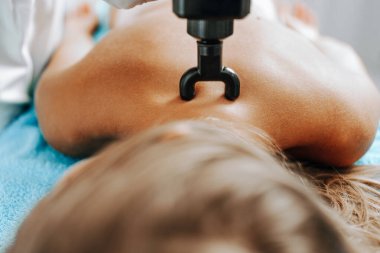 Young sporty Caucasian woman having body massage using percussion pistol at beauty relax center. Electric massager pistol for special muscle treatment. Sports and pain recovery concept.