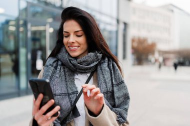 Şehir caddesinde dikilen güzel modaya uygun bayan turist çevrimiçi iletişim ve sosyal medya içeriği oluşturmak için elektronik akıllı telefon kullanıyor..